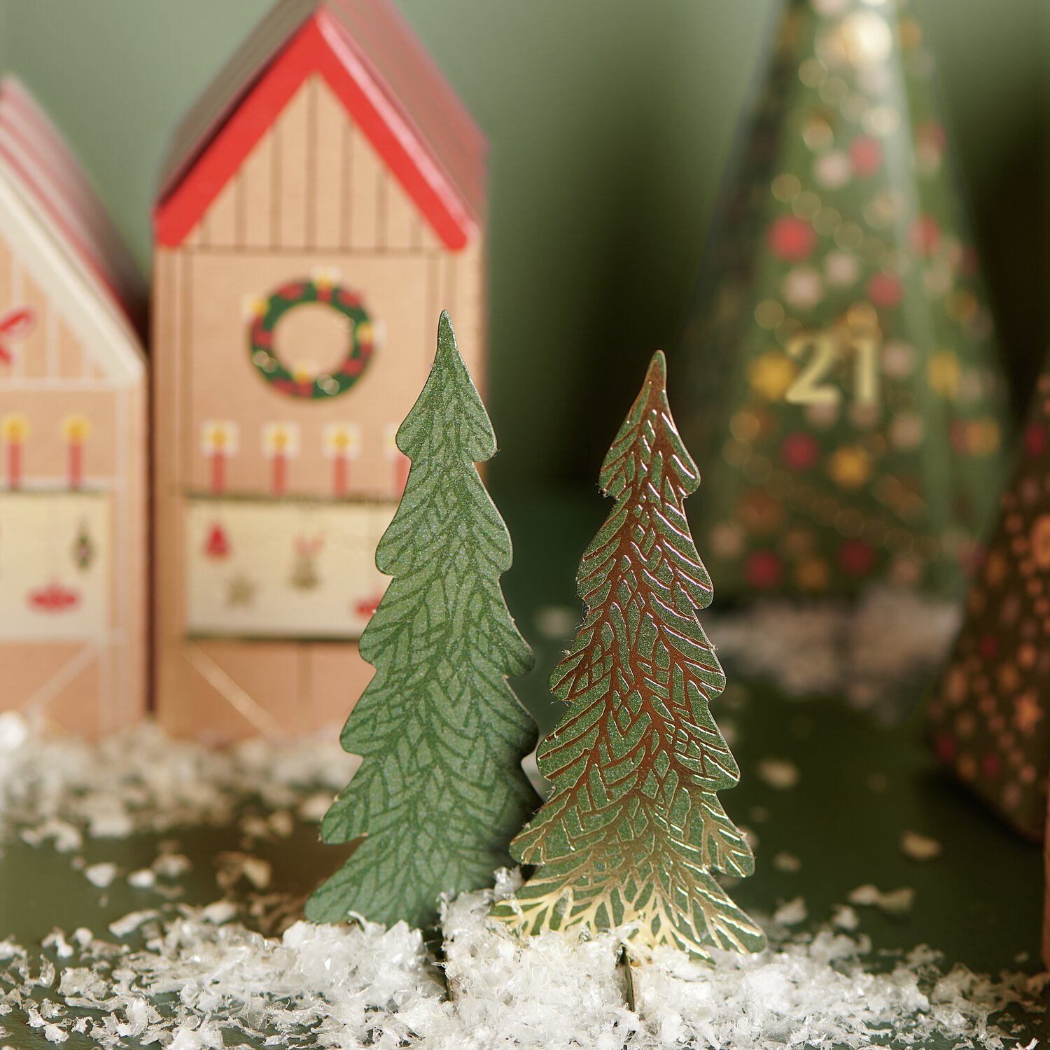 Adventskalender Bastelset mit grünen Tannenbäumen und dekorativen Häuschen im Hintergrund.