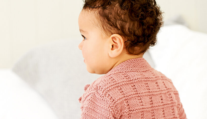 Strickanleitung Babyjacke mit Rundhalsausschnitt, Baby von hinten mit strukturierter, rosafarbener Strickjacke, weicher Hintergrund im Kinderzimmer