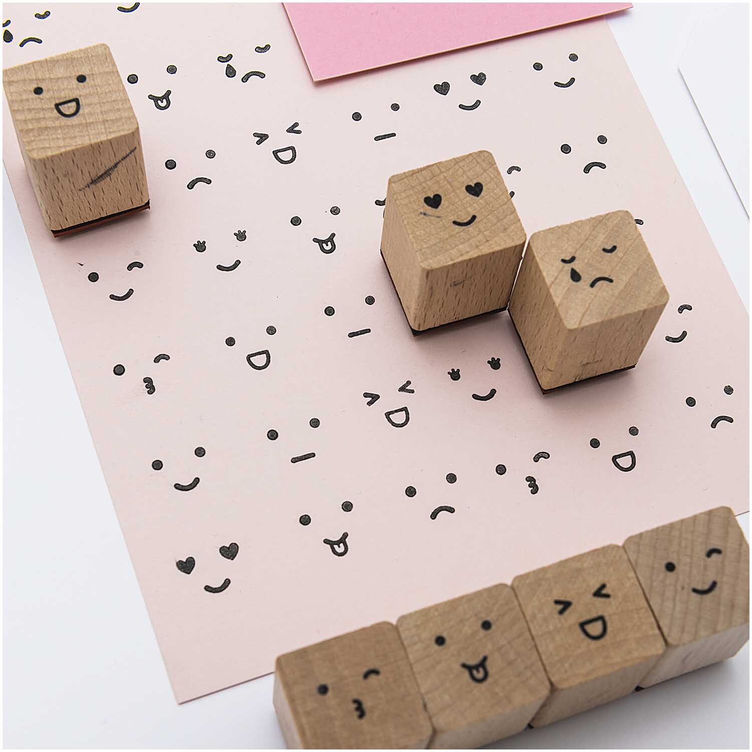 Paper Poetry Stempelset Smile, 13-teilig, von Rico Design. Holzstempel mit verschiedenen Gesichtsausdrücken auf rosa Papier.