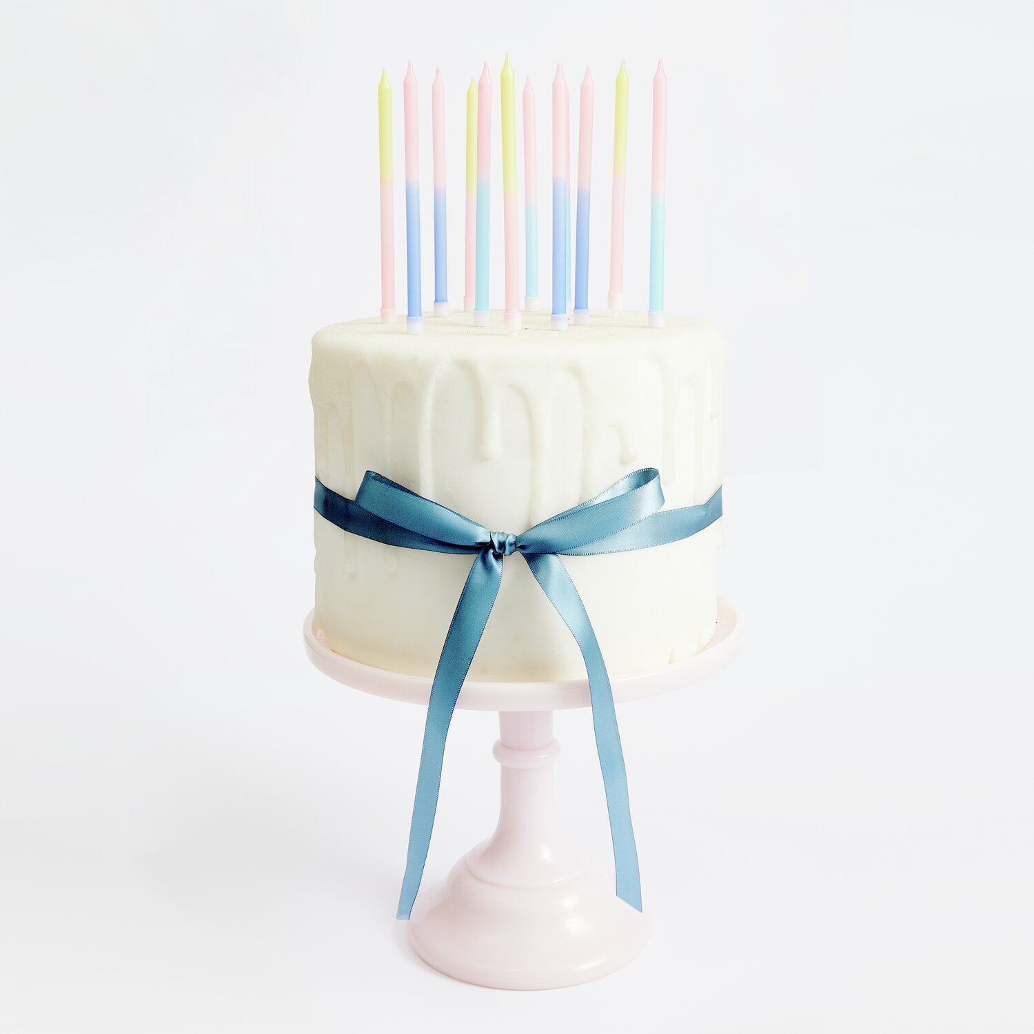 Kerzen Dip Dye Blau/Pink/Gelb auf weißem Kuchen mit blauer Schleife, präsentiert auf rosa Tortenständer.