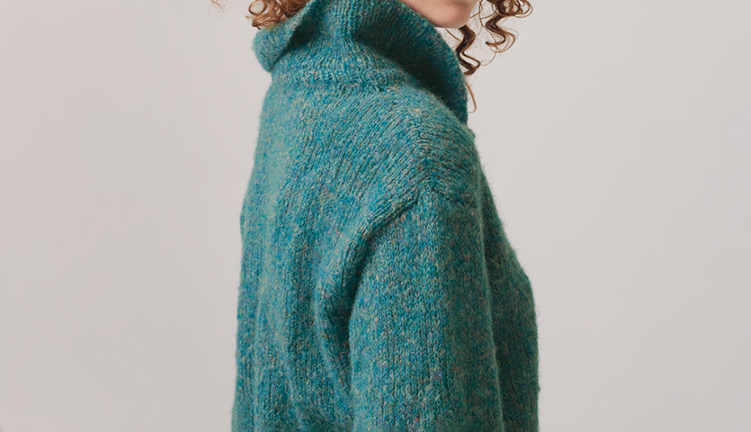 Strickanleitung Kragen-Pulli aus Fashion Skinny Alpaca aran: Frau trägt türkisfarbenen Pullover mit hohem Kragen, seitlich fotografiert.
