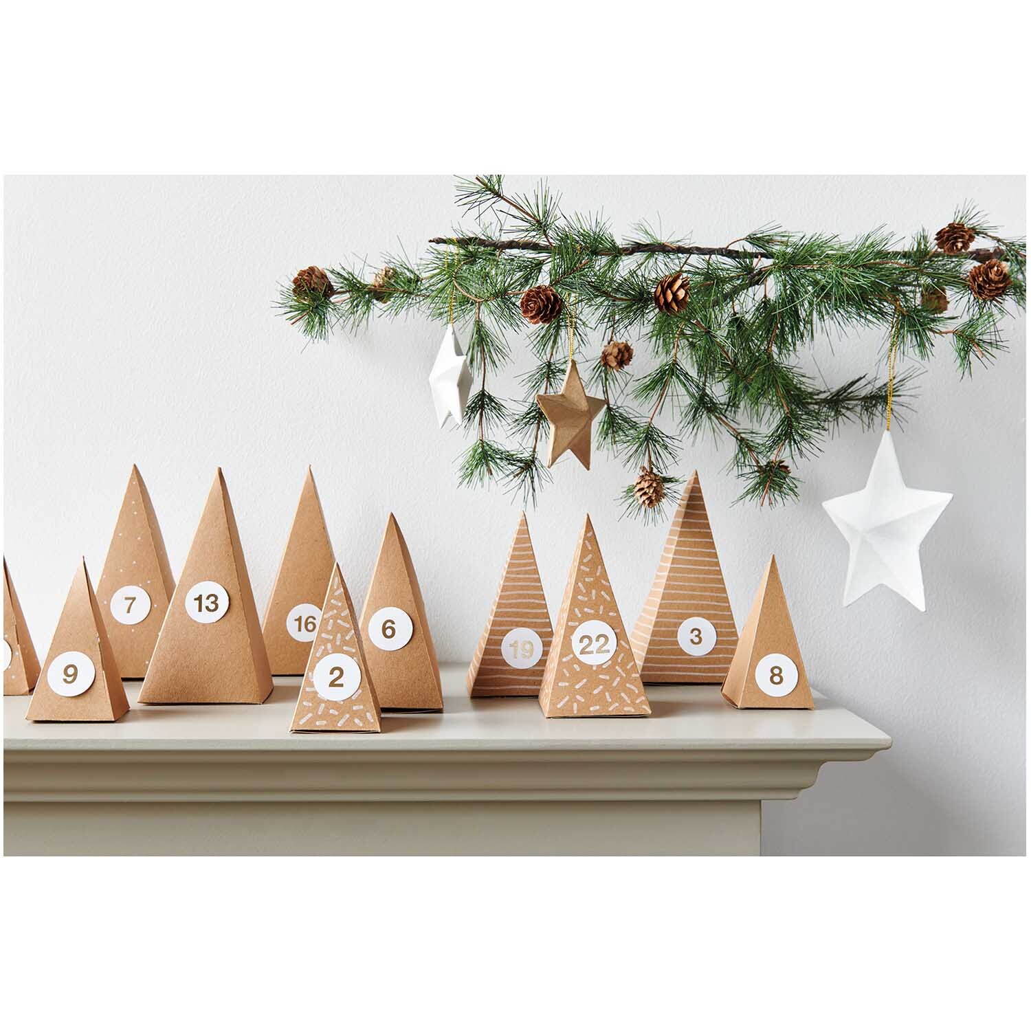 Paper Poetry Adventskalender Bäume 24 Stück aus Kraftpapier auf einem Regal, dekoriert mit Tannenzweigen und Sternen.
