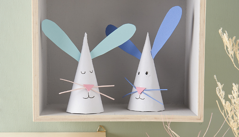Bastelanleitung Osterhasen aus Spitztüten: Zwei weiße Hasen mit blauen und grünen Ohren in einem Regal.