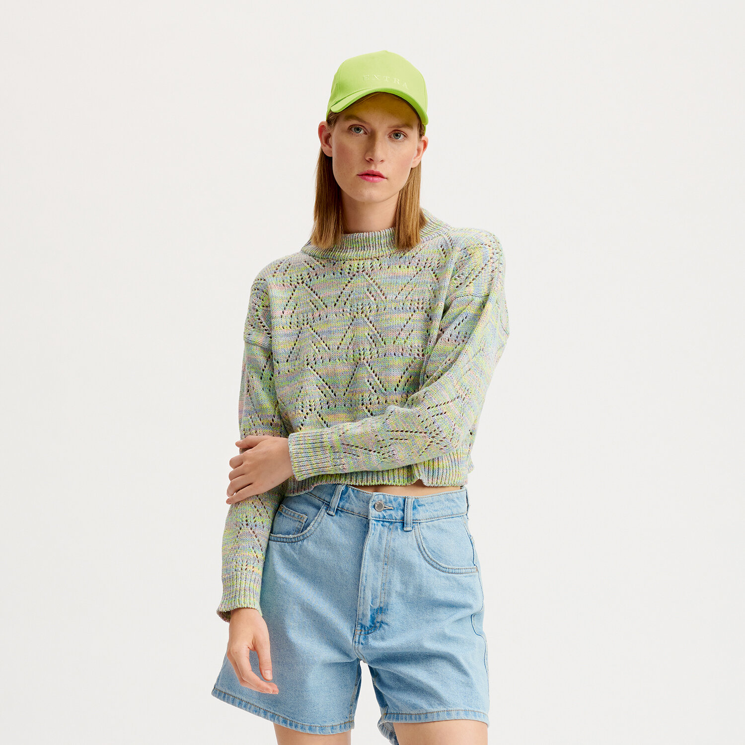 Strickanleitung Pullover aus Creative Summer Sprinkles: Frau trägt gestrickten Pullover in Pastellfarben, kombiniert mit blauer Jeansshorts und grüner Kappe.