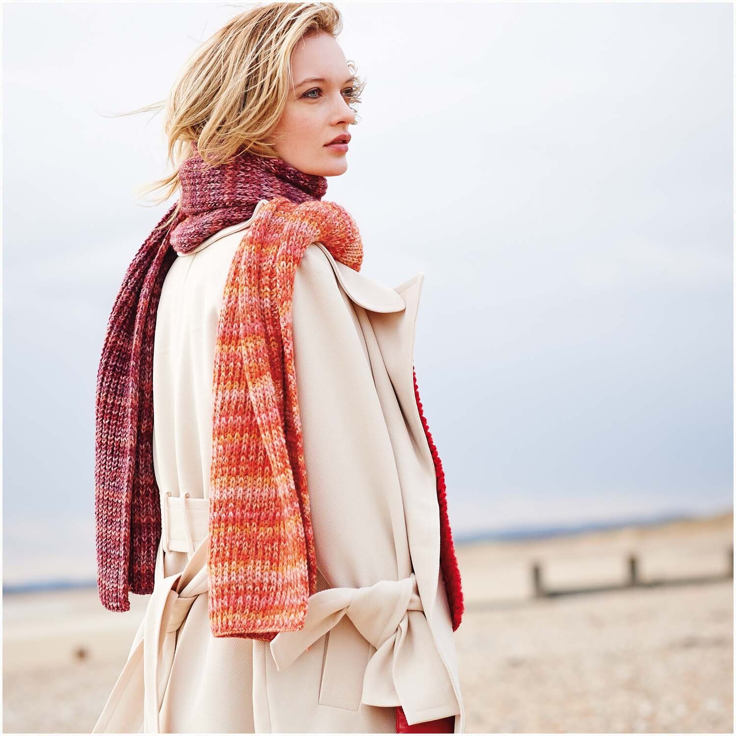 Essentials Alpaca Blend Print Chunky: Frau trägt gestrickten Schal in Rot-Orange und Lila-Rot von Rico Design, passend zu beigem Mantel.
