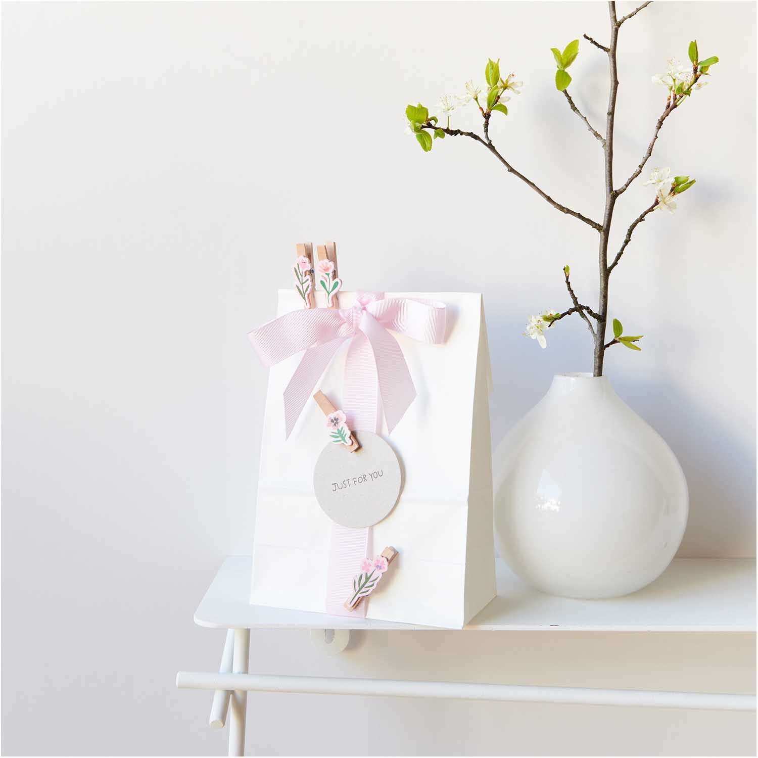 Dekoklammern Blumen Mix natur 4 Stück von Rico Design: Mehrfarbige Klammern verzieren eine weiße Geschenktüte mit rosa Schleife, daneben eine Vase mit Zweig.