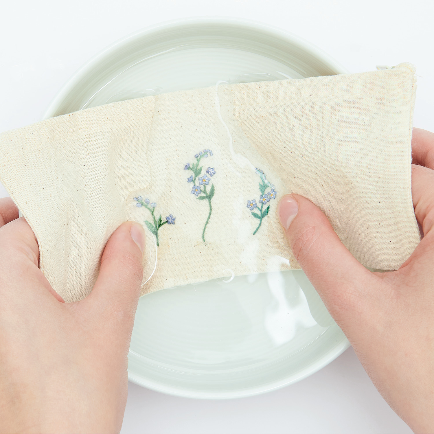Stick and Stitch Blümchen: Hände halten ein besticktes Tuch mit Blumenmotiv über einer Schüssel. Ideal für kreative Projekte.