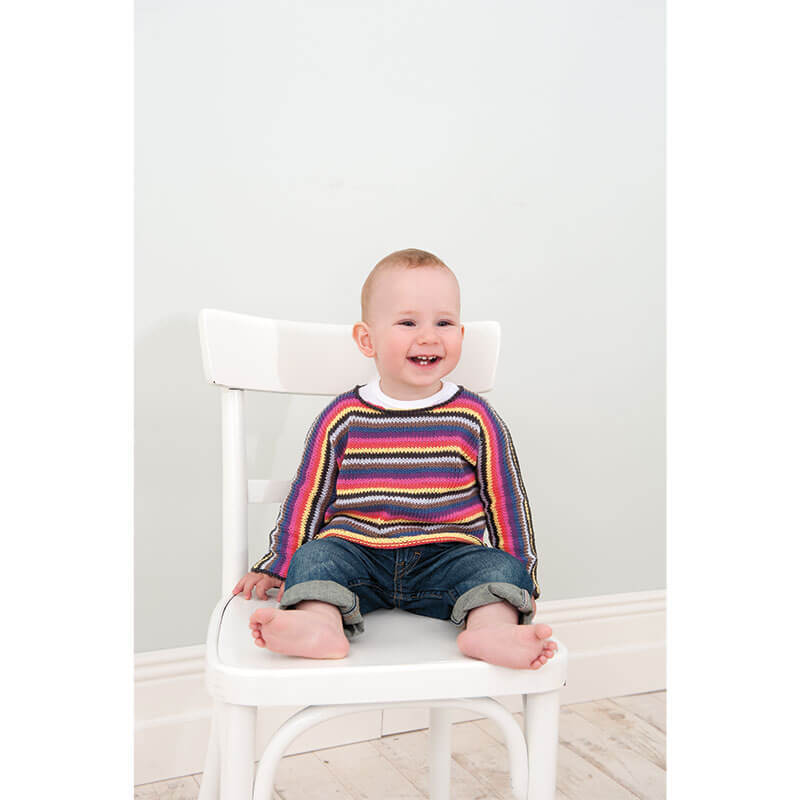 Rico Baby Heft Nr.9: Ein lächelndes Baby in einem bunten Strickpullover sitzt auf einem weißen Stuhl.