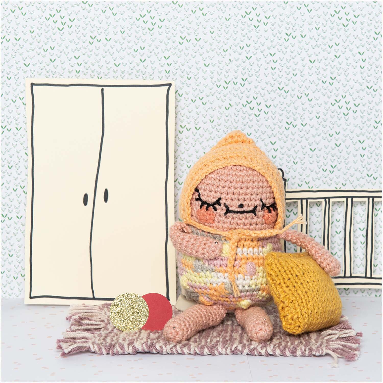 Ricorumi Crazy Cute Family deutsch: Häkelfigur in buntem Outfit sitzt auf Teppich mit gelbem Kissen, vor gezeichneter Wand.