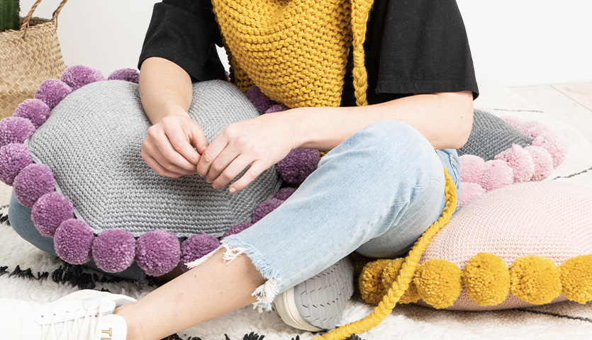 Häkelanleitung Kissen aus Essentials Mega Wool chunky: Person sitzt auf bunten Kissen mit Pompons.