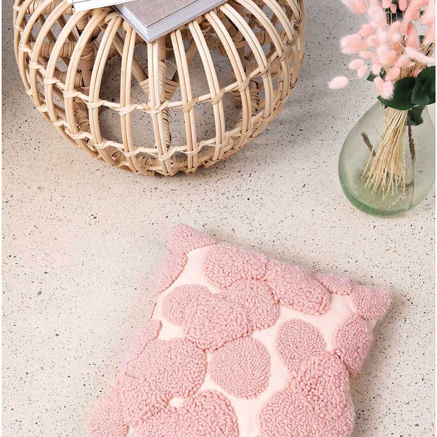 Punch Needle Packung Kissen Punkte rosa von Rico Design auf einem hellen Teppich neben einem runden Rattanhocker und einer Vase mit Trockenblumen.