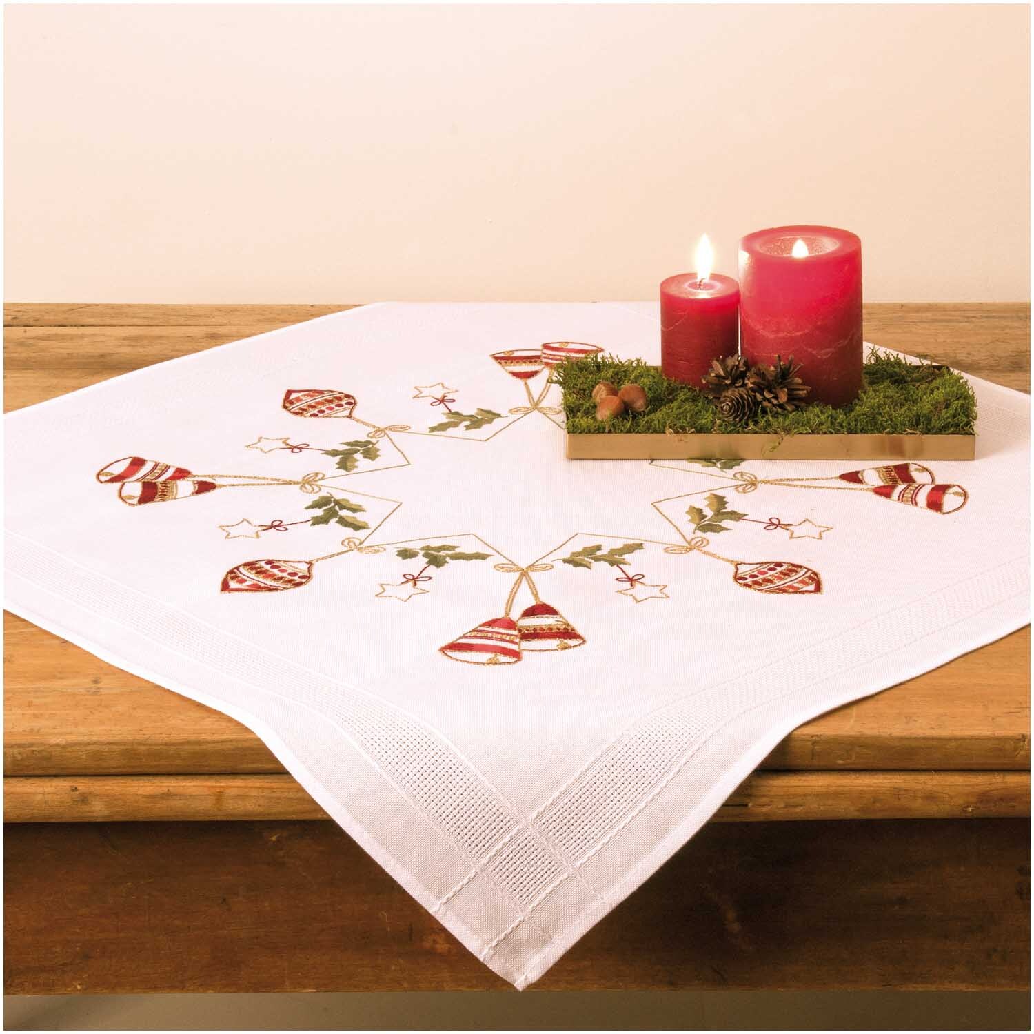 Stickpackung Tischdecke Stern mit Kugeln 80x80cm zeigt festliche Stickerei mit roten Kerzen und Tannenzweigen auf einem Holztisch.