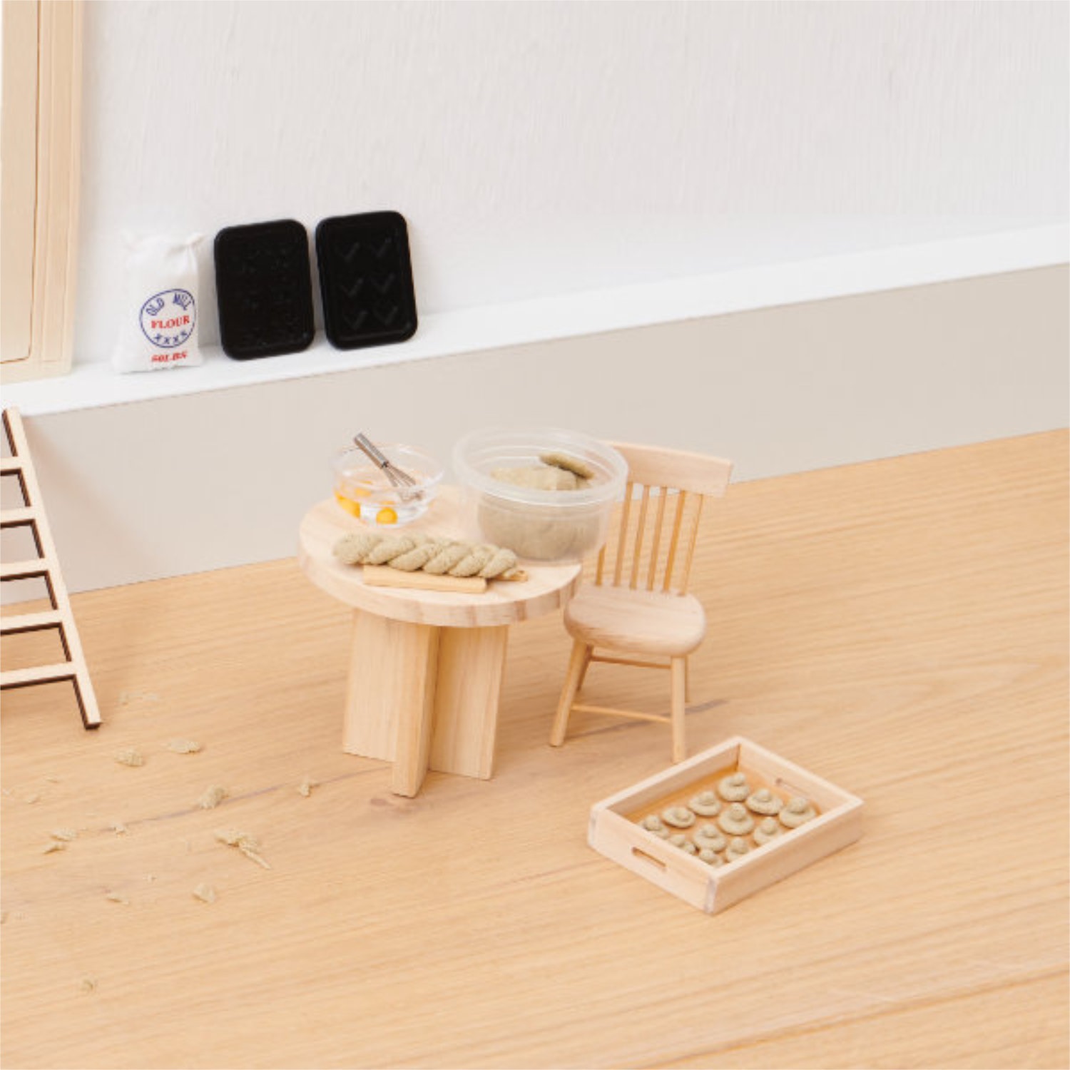 Miniatur Tisch und Stuhl mit zwei Backschüsseln, einem Tablett, Backbleche, eine Packung Mehl und Miniatur-Hefezopf.