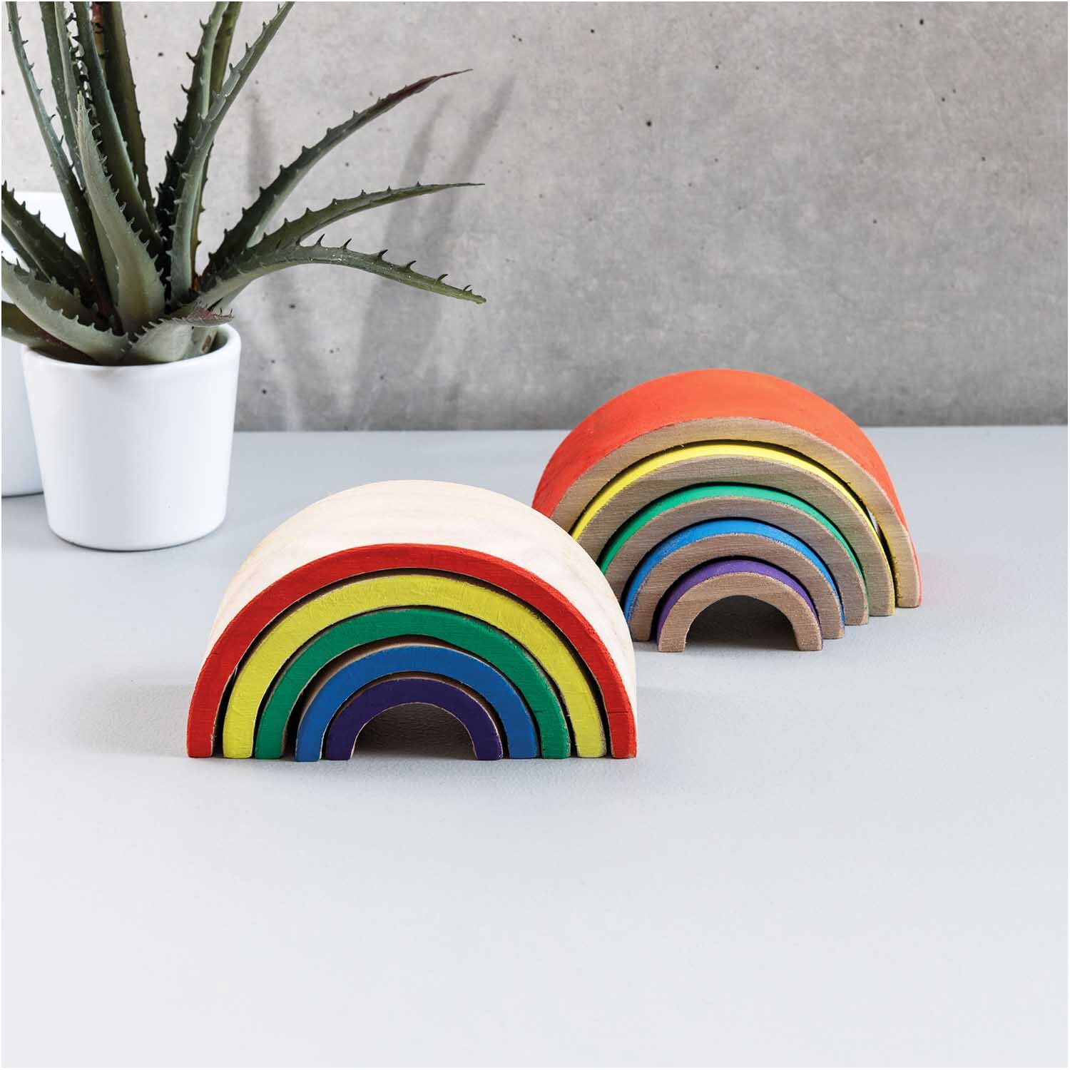 Holz-Regenbogen 12x6x5cm 5teilig: Bunte, ineinander passende Holzbögen vor grauer Wand und Pflanze.