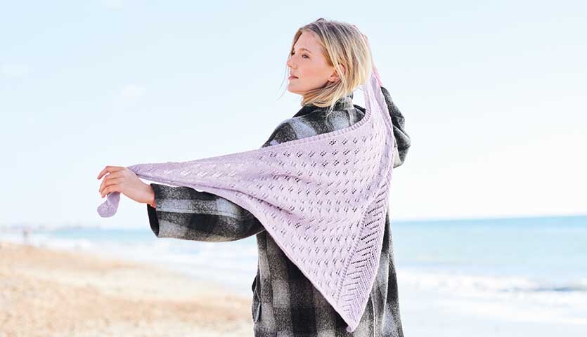 Strickanleitung Tuch aus Essentials Fine Fine Merino: Frau am Strand mit einem gestrickten, hellen Tuch über den Schultern, das sanft im Wind weht.