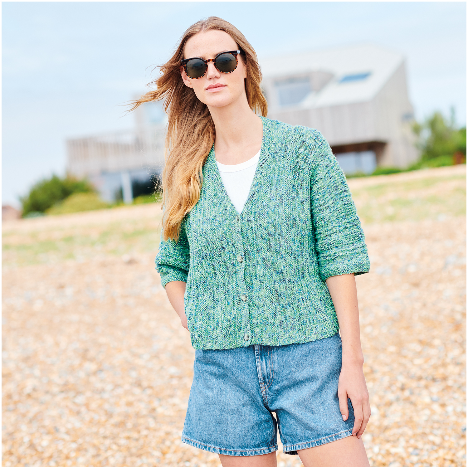 Strickset Jacke Modell 24 aus Lovewool Nr. 18: Frau am Strand in grüner Strickjacke und Jeansshorts.