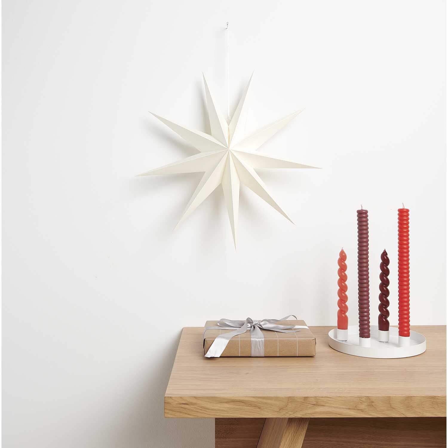Papierstern auffaltbar 45cm von Rico Design an einer weißen Wand, neben Kerzen und Geschenk auf Holztisch.