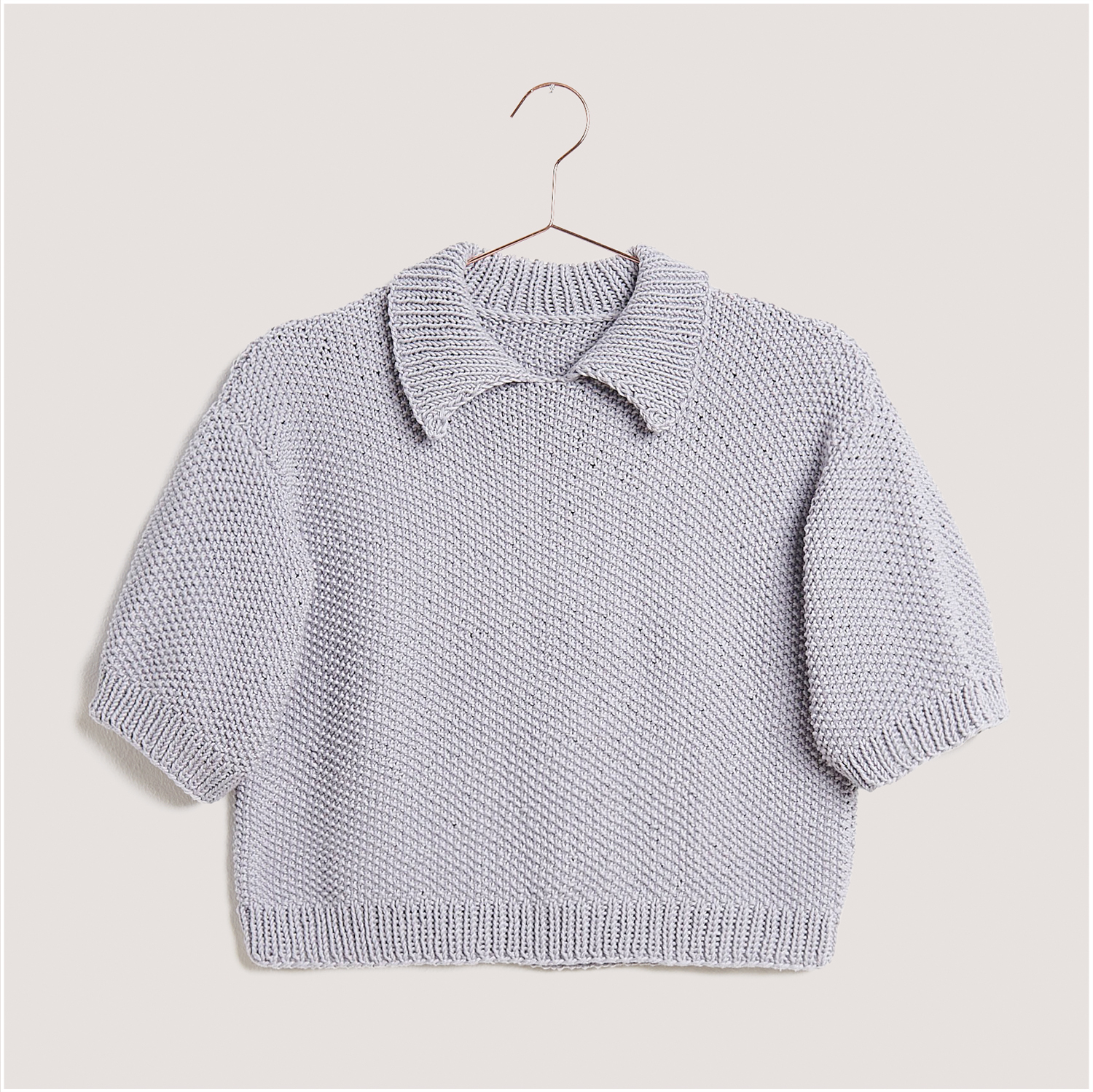 Strickset Shirt Modell 11 aus Lovewool Nr. 18: Ein graues, kurzärmeliges Strickshirt mit Kragen, hängend an einem Kleiderbügel.