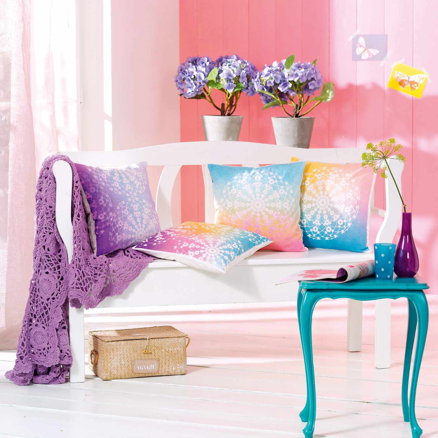 Fashion Spray: Bunte Kissen auf weißer Bank mit lila Decke, umgeben von blauen Blumen und einem türkisfarbenen Tisch.