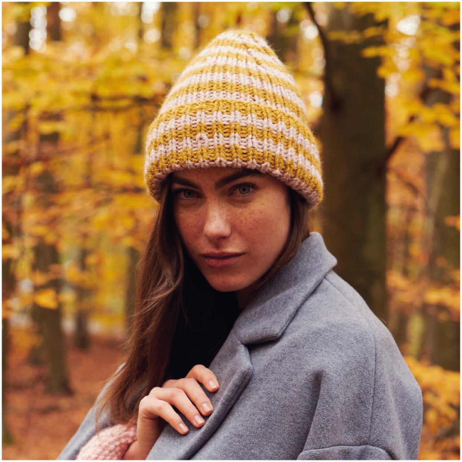 Essentials Alpaca Twist Chunky: Frau im Wald mit gestreifter Mütze in Karamell und Grau, Herbststimmung.
