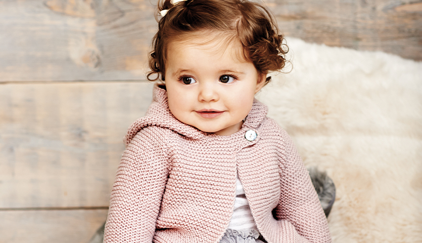 Strickanleitung Babyjacke mit Kapuze in Mausoptik: Niedliches Baby in rosa Strickjacke vor Holzhintergrund.