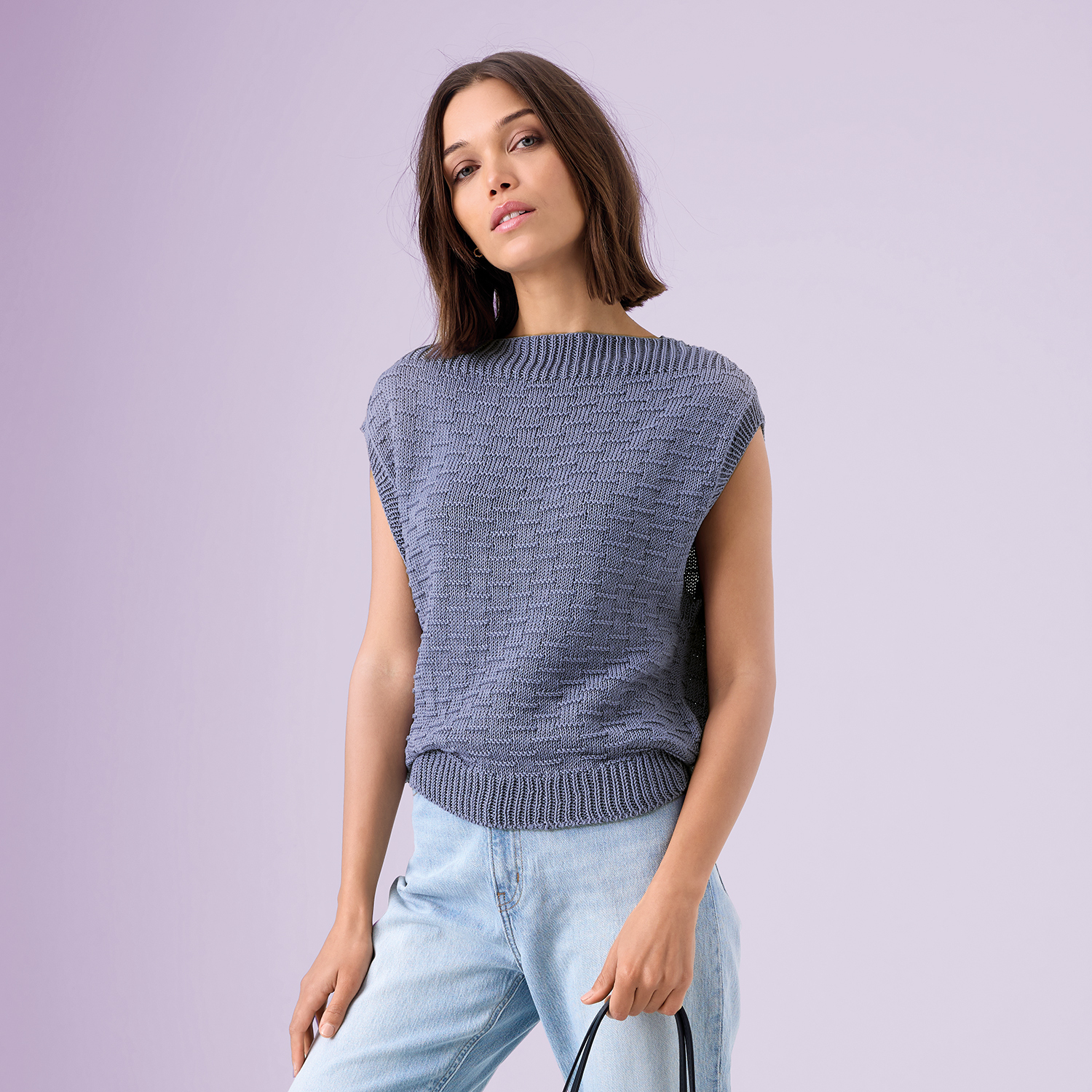 Strickanleitung Strukturmuster-Top aus Mille Fili: Frau trägt ein strukturiertes, ärmelloses Top in Blau, kombiniert mit hellen Jeans vor lila Hintergrund.