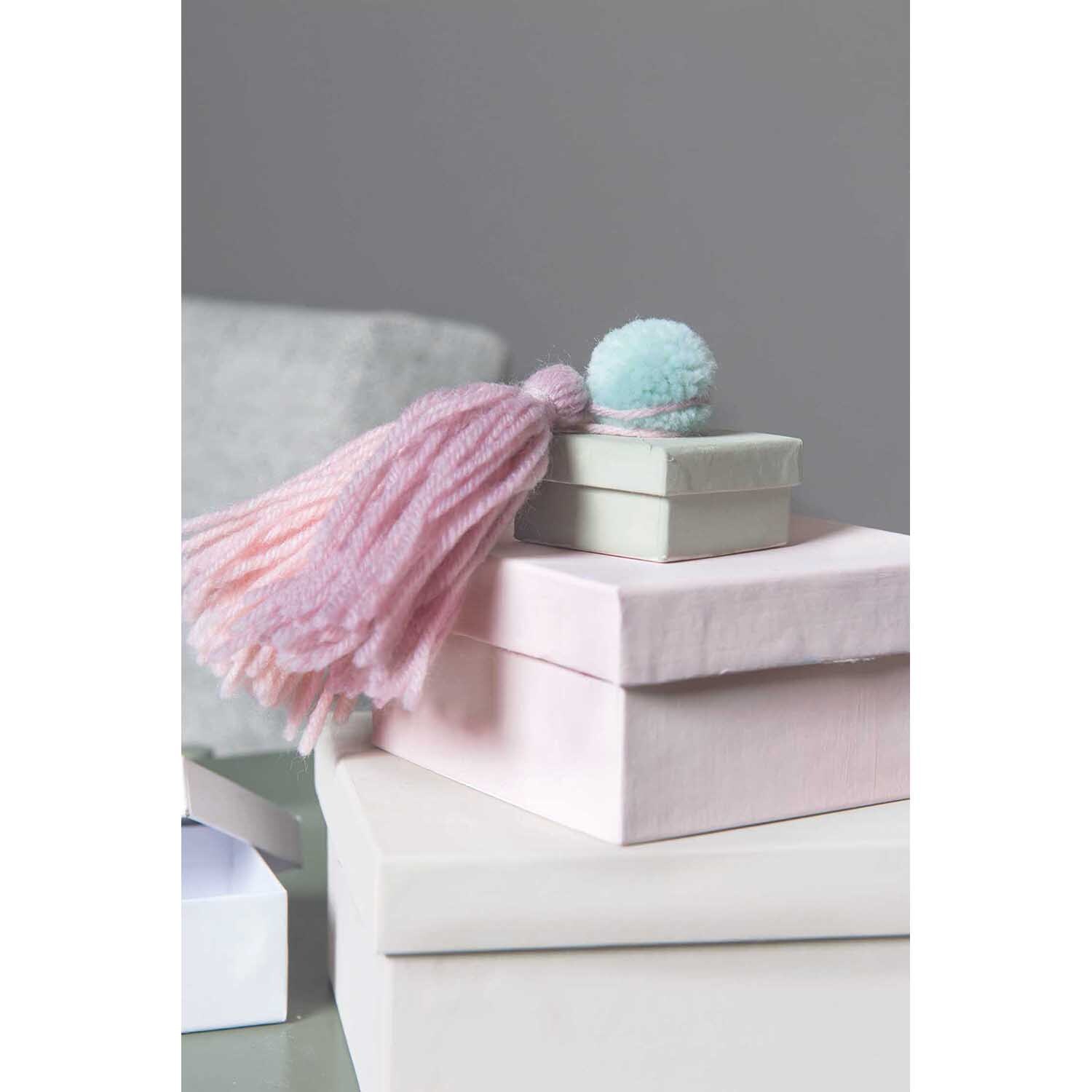 Garn Pompons 24 Stück von Rico Design auf gestapelten Geschenkboxen mit rosa Quaste und blauem Pompon.