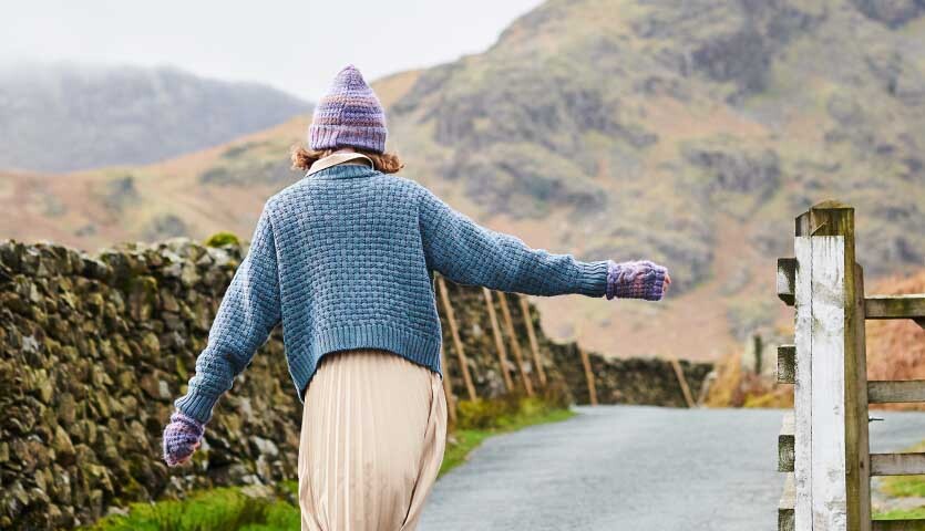 Strickanleitung Jacke aus Essentials Merino dk: Person in blauer Strickjacke und Mütze auf ländlichem Weg.