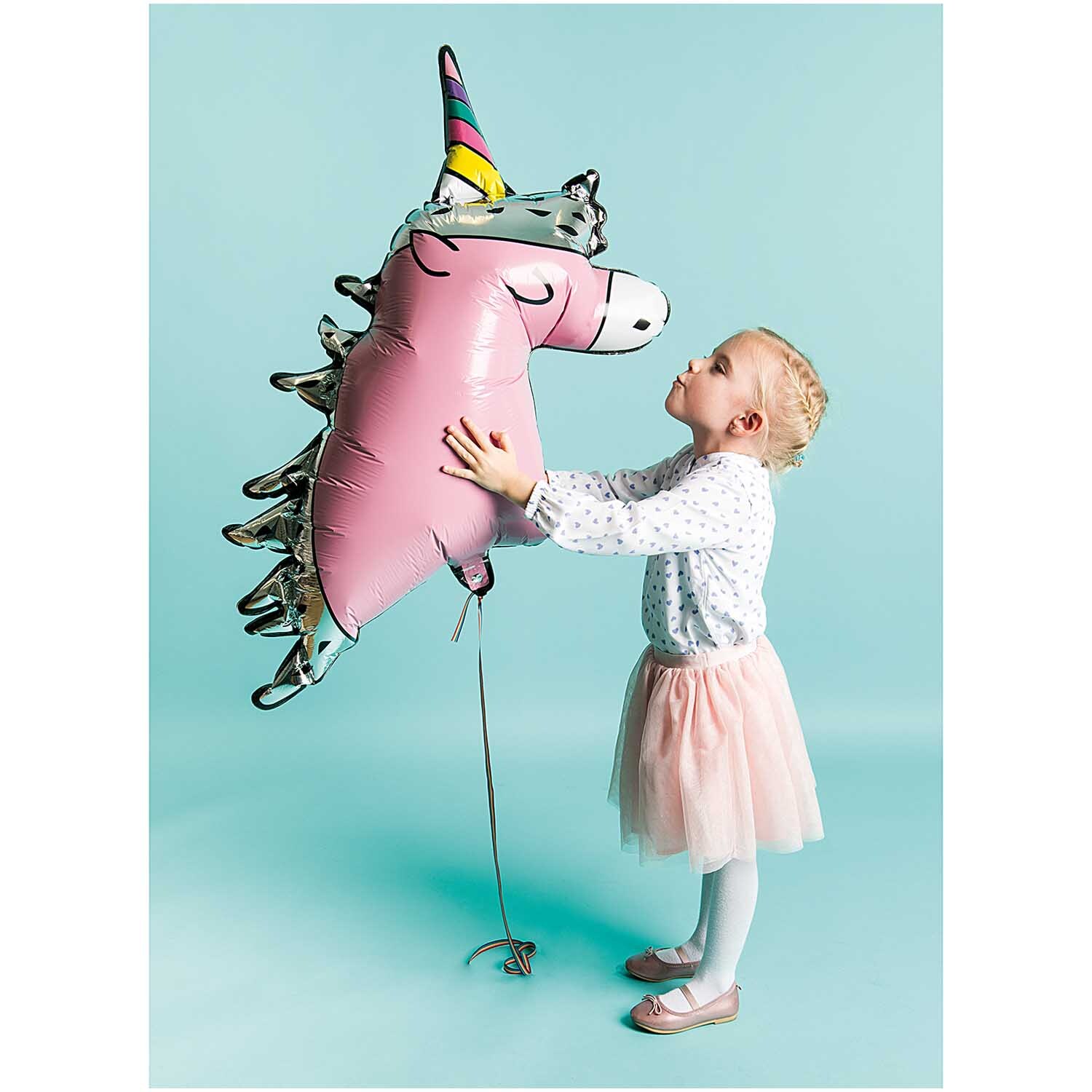 Folienballon Einhorn 85x83cm: Ein Mädchen umarmt einen rosa Einhornballon von Rico Design vor blauem Hintergrund.