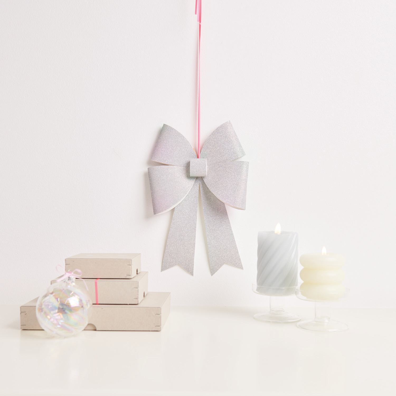 Paper Poetry Bastelset XL-Schleife Glitter, silber irisierend, hängt neben Kerzen und Geschenken.
