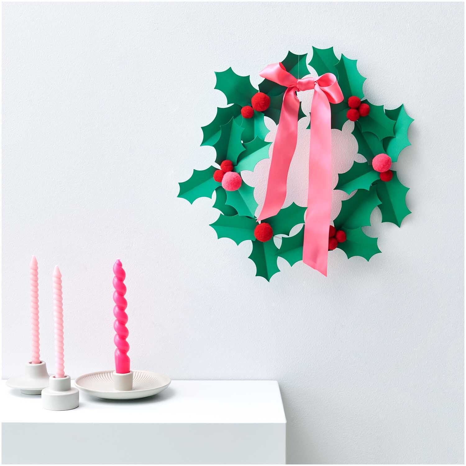 Paper Poetry Bastelset Ilex: Ein grüner Kranz mit roten Beeren und rosa Schleife, dekoriert neben rosa Kerzen.