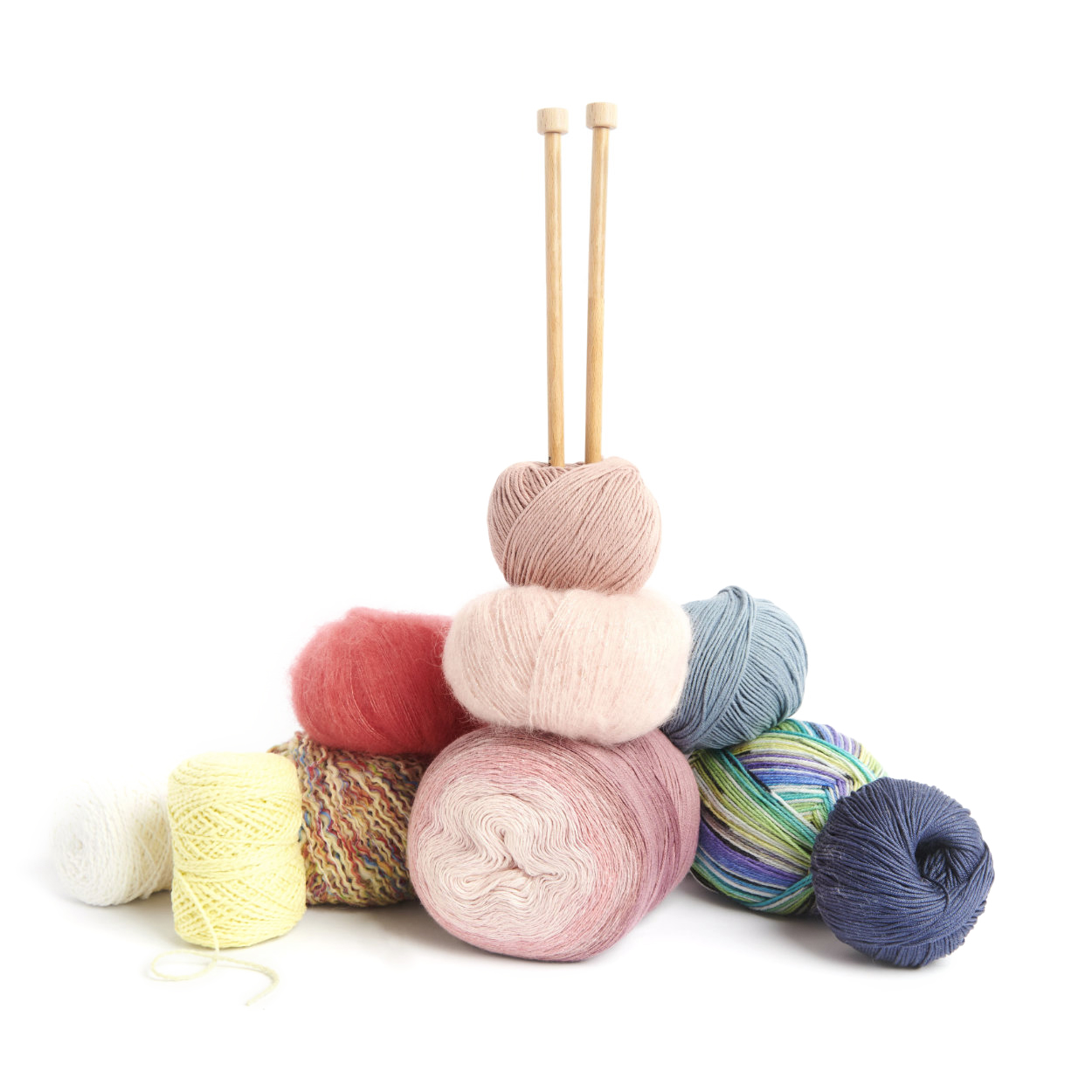 Verschiedene Garne liegen neben und übereinander und zwei Stricknadeln in ein Knäul gesteckt.