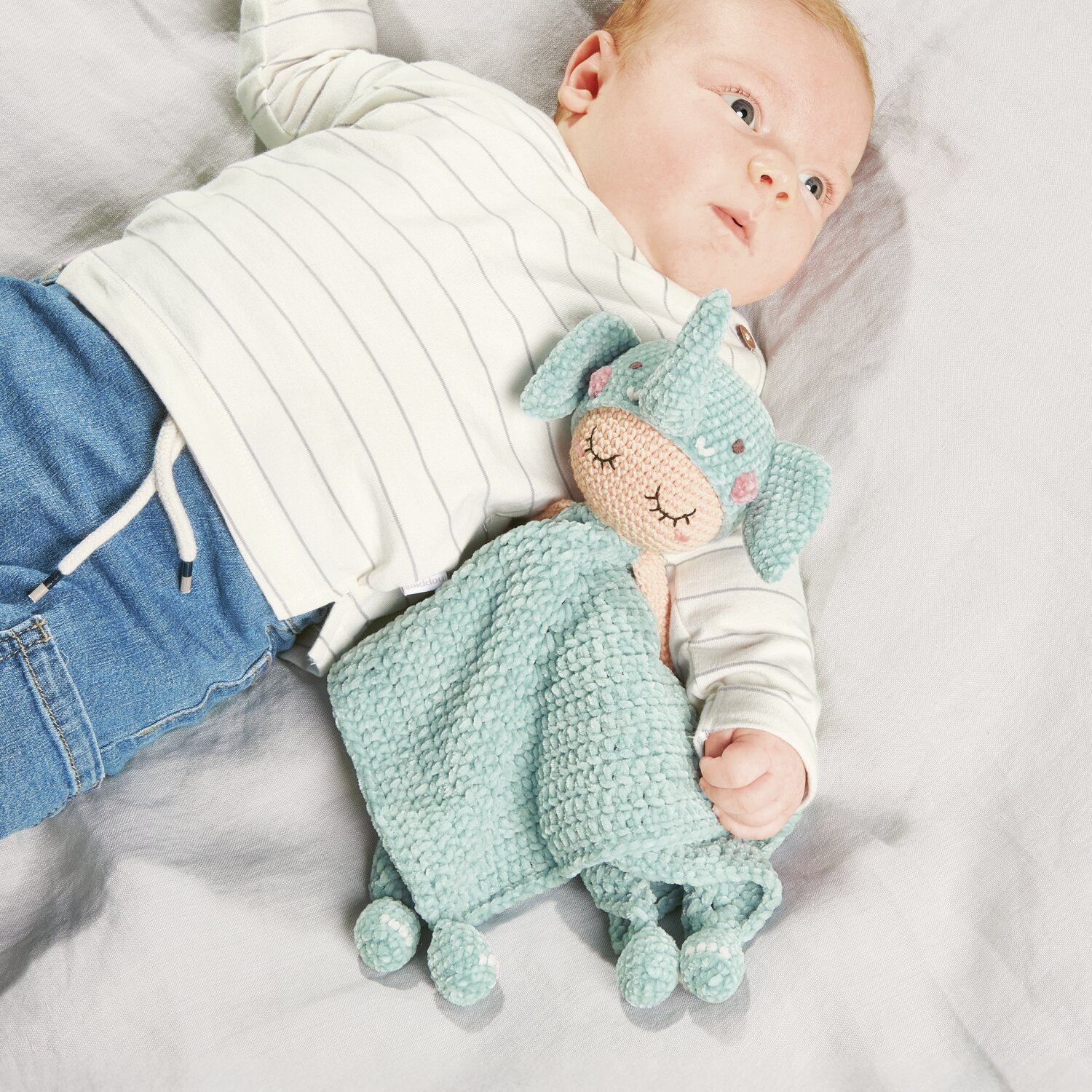 Ricorumi Baby Blankies Nilli Nilli: Ein Baby liegt auf einer Decke mit einem gehäkelten, aqua-farbenen Kuscheltuch von Rico Design.