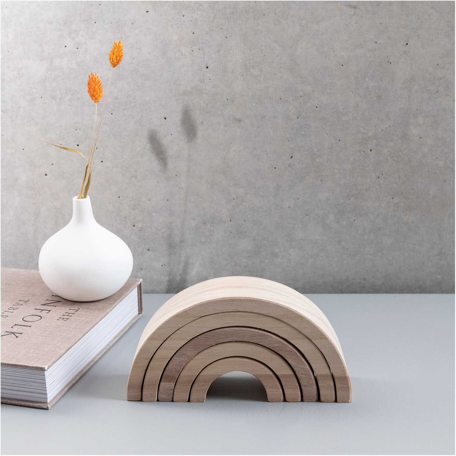 Holz-Regenbogen 18x9x5cm 5teilig: Dekoratives Holzspielzeug in Naturtönen vor grauer Wand, neben Vase und Buch.