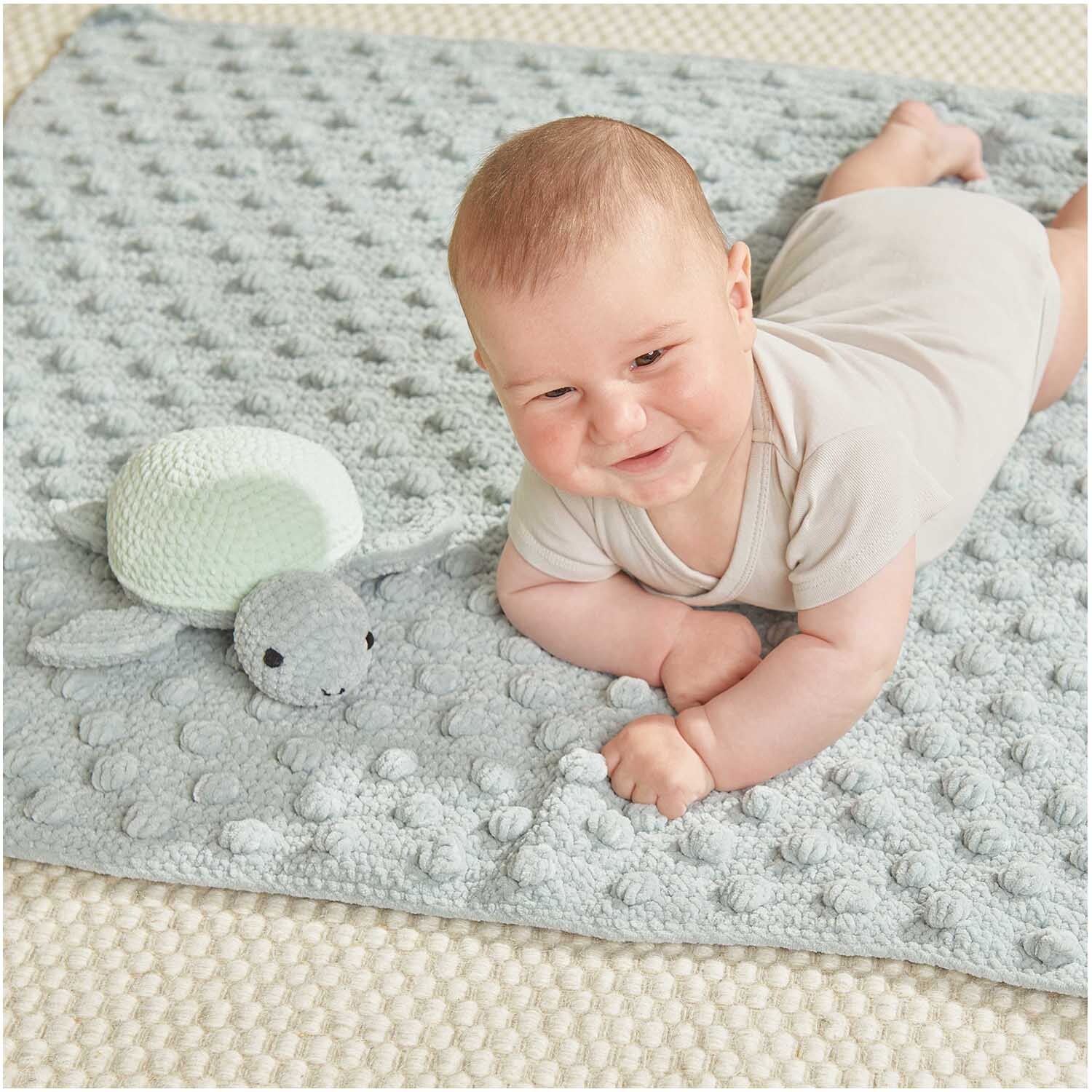 Creative Chenillove: Ein Baby liegt auf einer weichen, hellblauen Decke mit einer gestrickten Schildkröte.