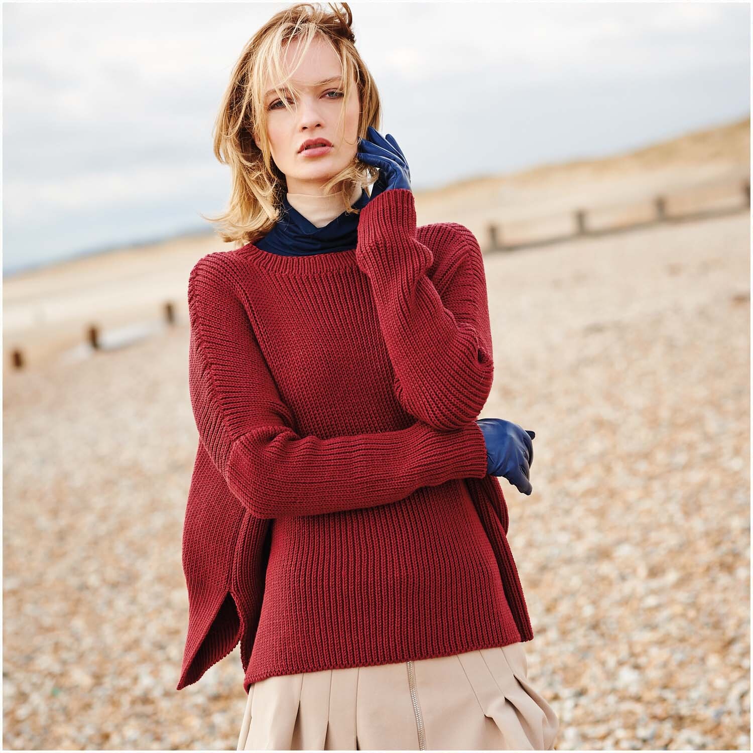 Essentials Merino dk: Frau am Strand in rotem Pullover, beige Rock, blauer Schal, Rico Design.