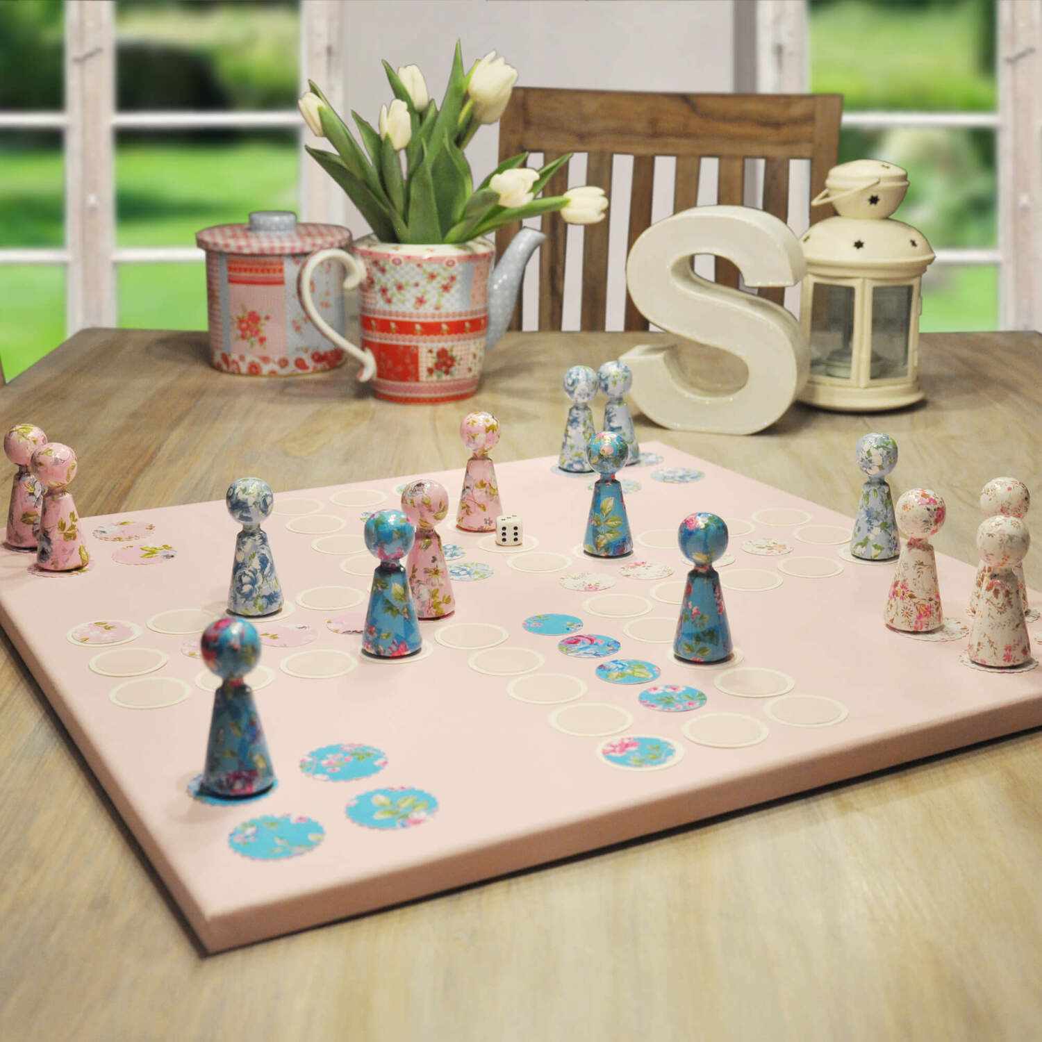 Bastelanleitung Brettspiel: Ein handgefertigtes Brettspiel mit bunten Spielfiguren auf einem Tisch. Im Hintergrund sind Blumen und Dekorationen zu sehen.