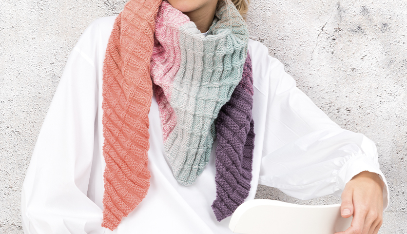 Strickanleitung Dreieckstuch aus Creative Wool Dégradé Super 6: Frau trägt farbenfrohes, gestreiftes Tuch in Rosa, Blau und Lila vor grauem Hintergrund.