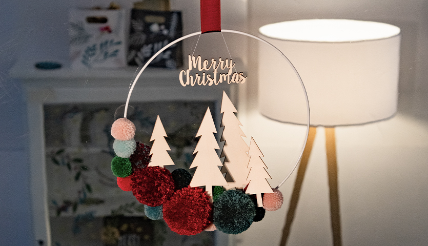 Bastelanleitung weihnachtlicher Pompon-Kranz mit roten und grünen Pompons, Holzbäumen und "Merry Christmas"-Schriftzug, vor einer Lampe.