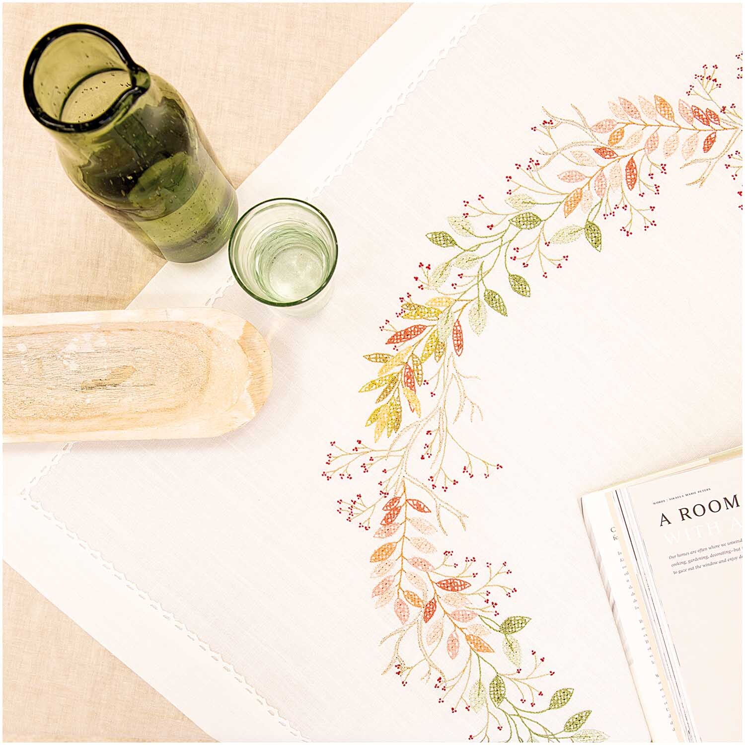 Stickpackung Tischdecke Herbstkranz 90x90cm zeigt ein herbstliches Blattdesign auf weißem Stoff, dekoriert mit Glasflaschen und Holzbrett.