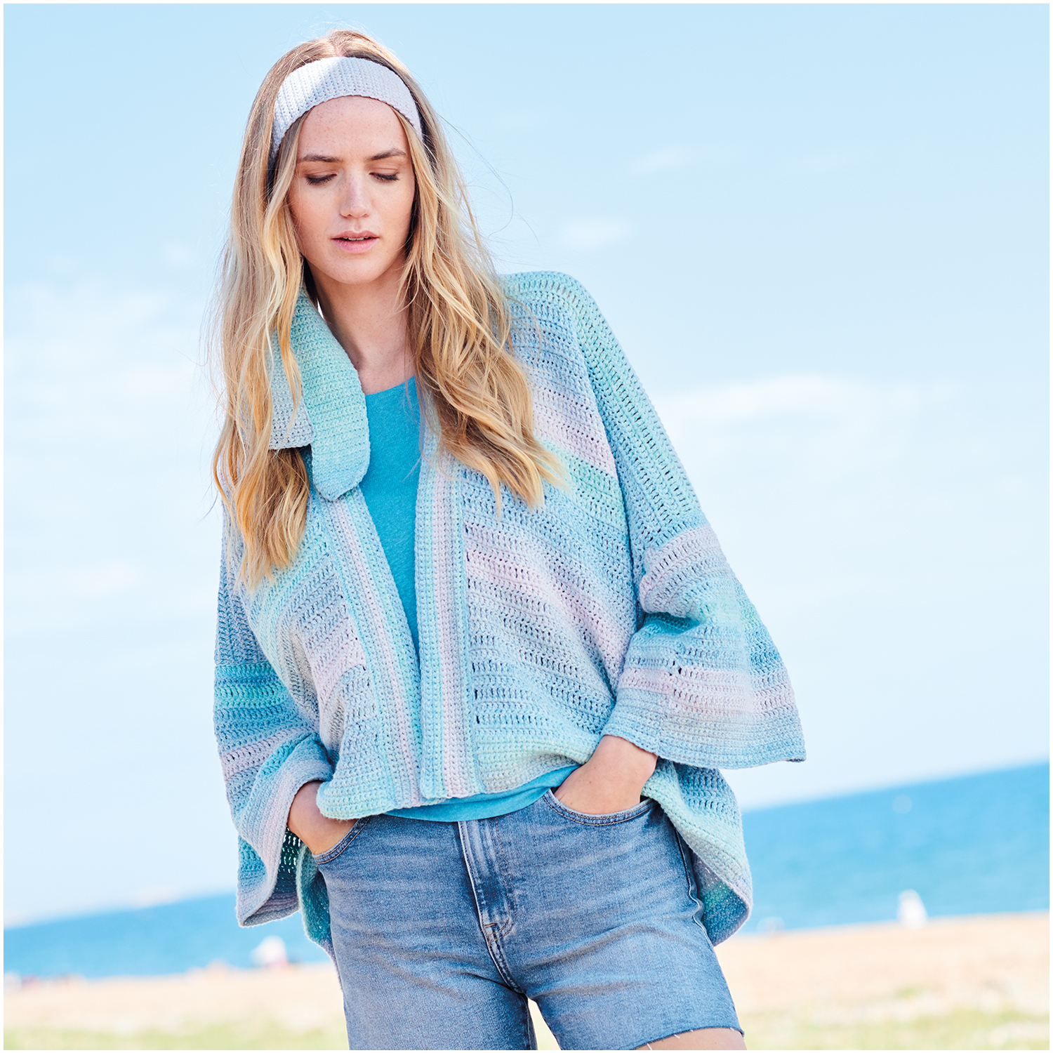 Häkelset Kimono-Jacke Modell 29: Frau am Strand trägt pastellfarbene Jacke und Jeansshorts.