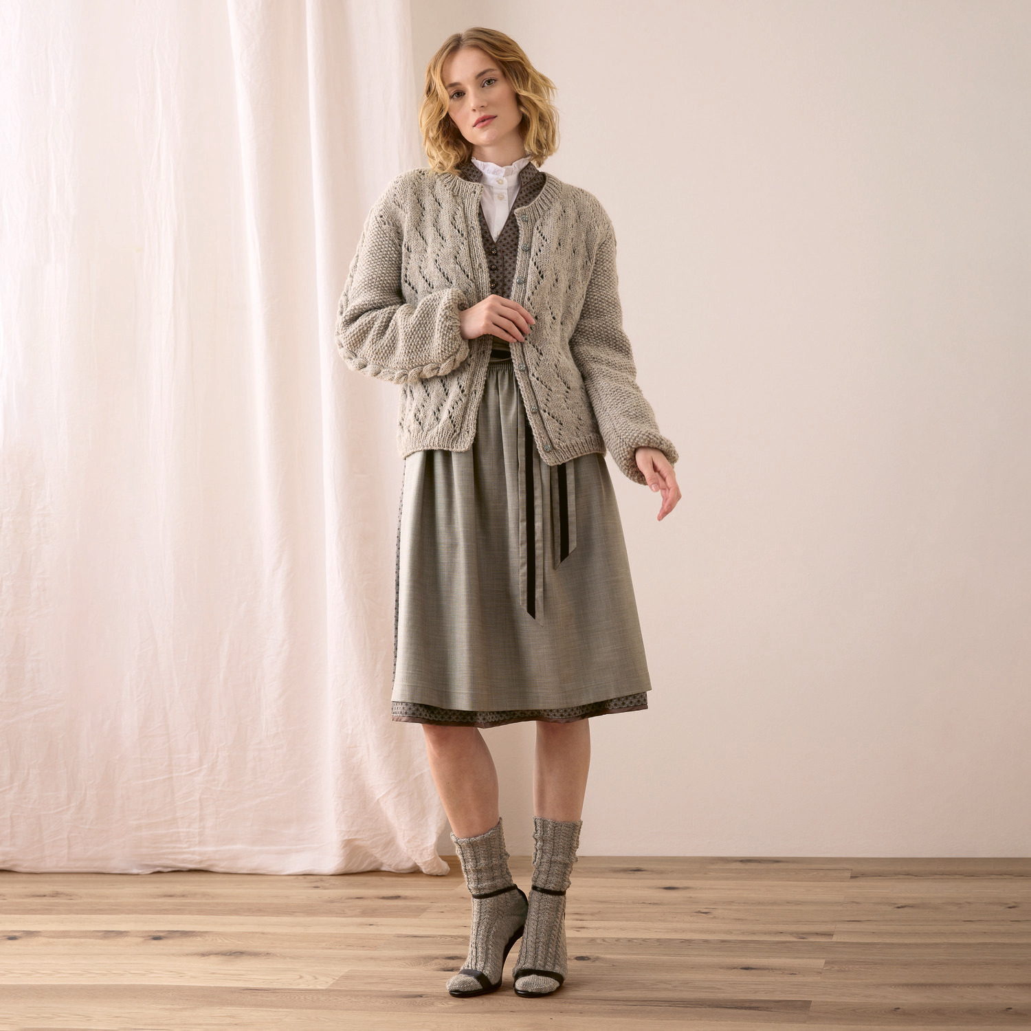 Strickset Jacke Modell 01c aus Trachtenbuch I mog di: Frau in grauer Strickjacke und Kleid, stehend auf Holzboden vor hellem Vorhang.