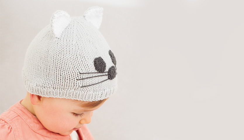 Strickanleitung Babymütze mit Katzenohren: Baby trägt eine graue Mütze mit niedlichen Katzenohren und Gesichtsmuster.