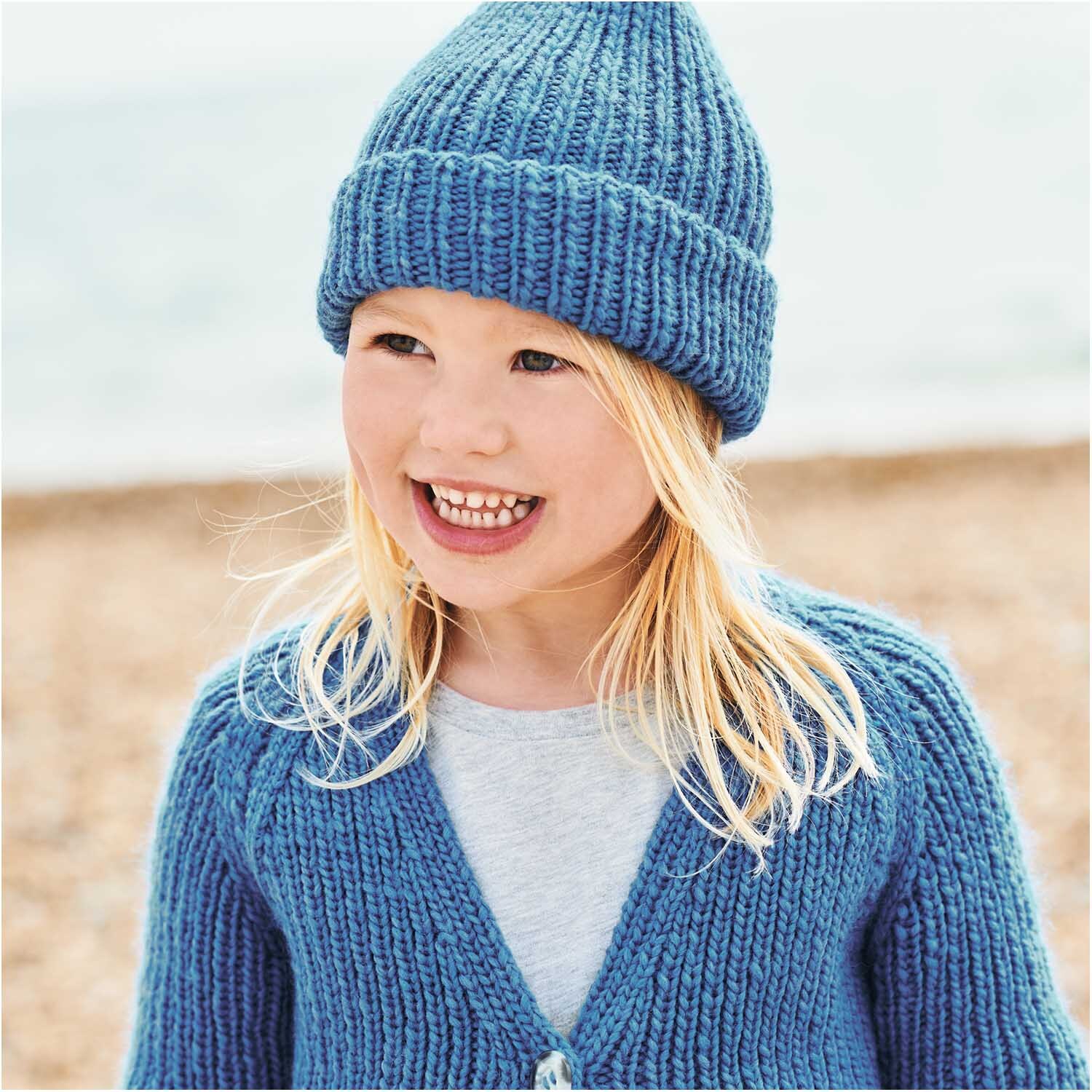 Essentials Super aran: Kind in gestricktem, azurblauem Outfit von Rico Design am Strand.
