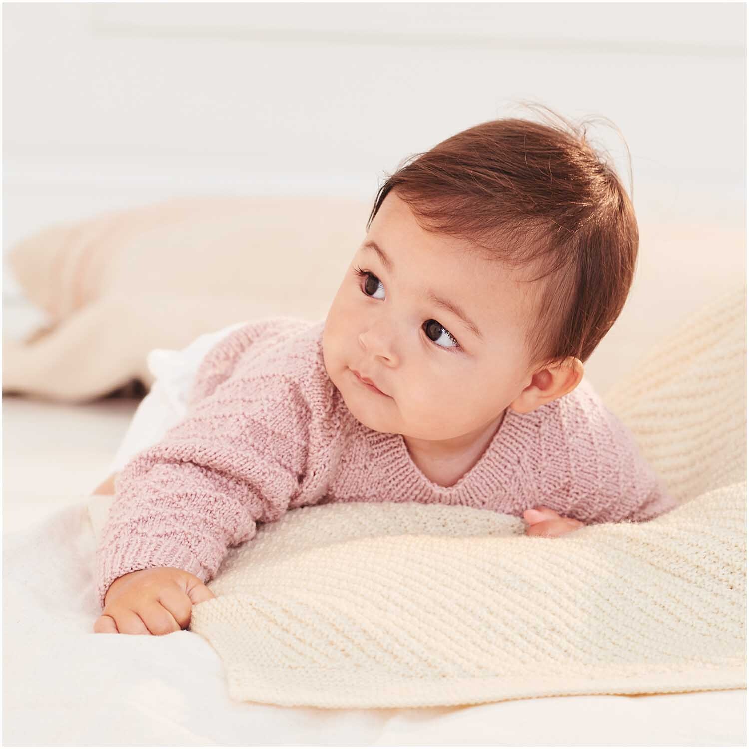 Baby Merino: Ein Baby in einem weichen, rosa Strickpullover von Rico Design liegt auf einer cremefarbenen Decke.