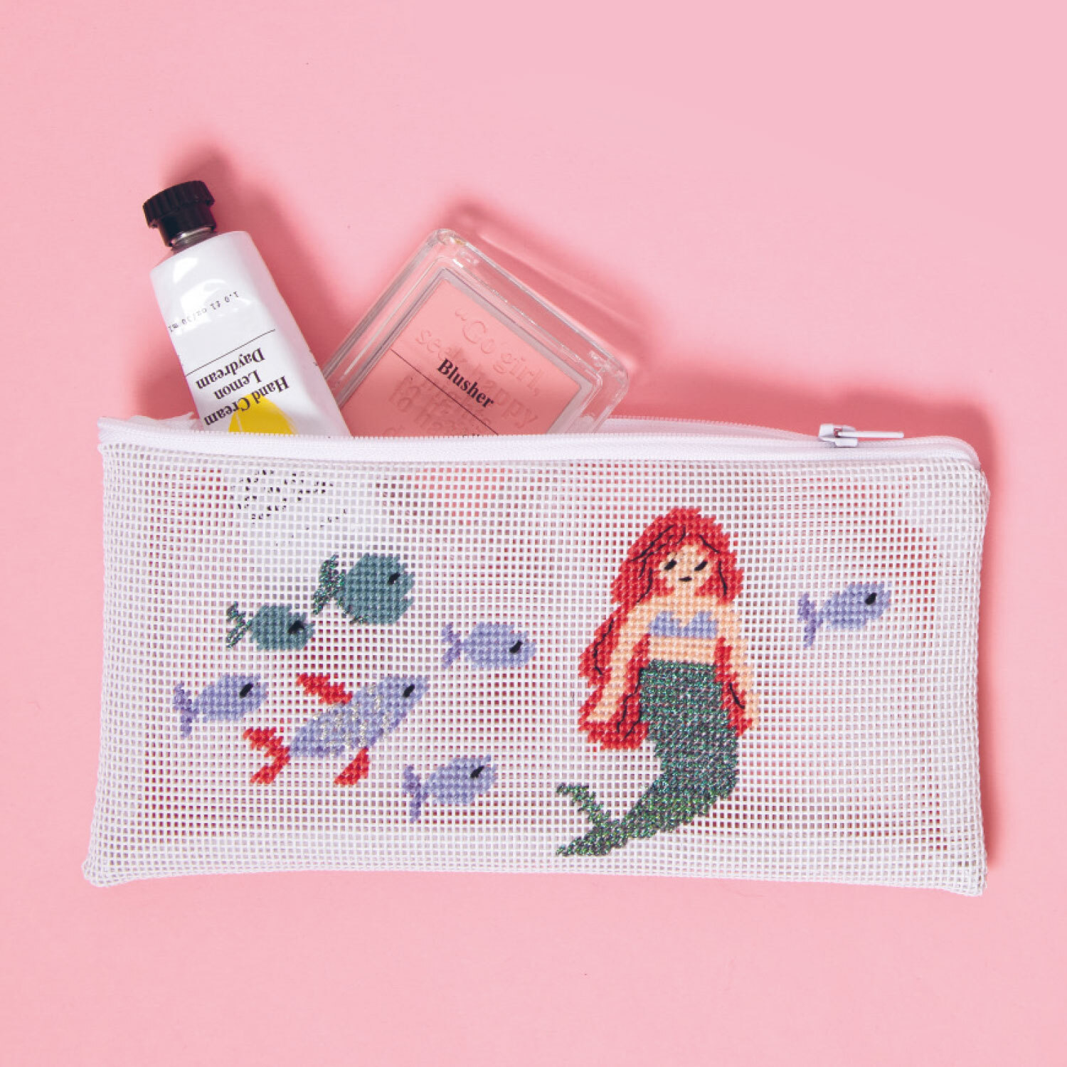 Stickanleitung Meerjungfrau Etui gestalten: Ein weißes Etui mit Meerjungfrau-Stickerei, Fischen und Kosmetik auf rosa Hintergrund.