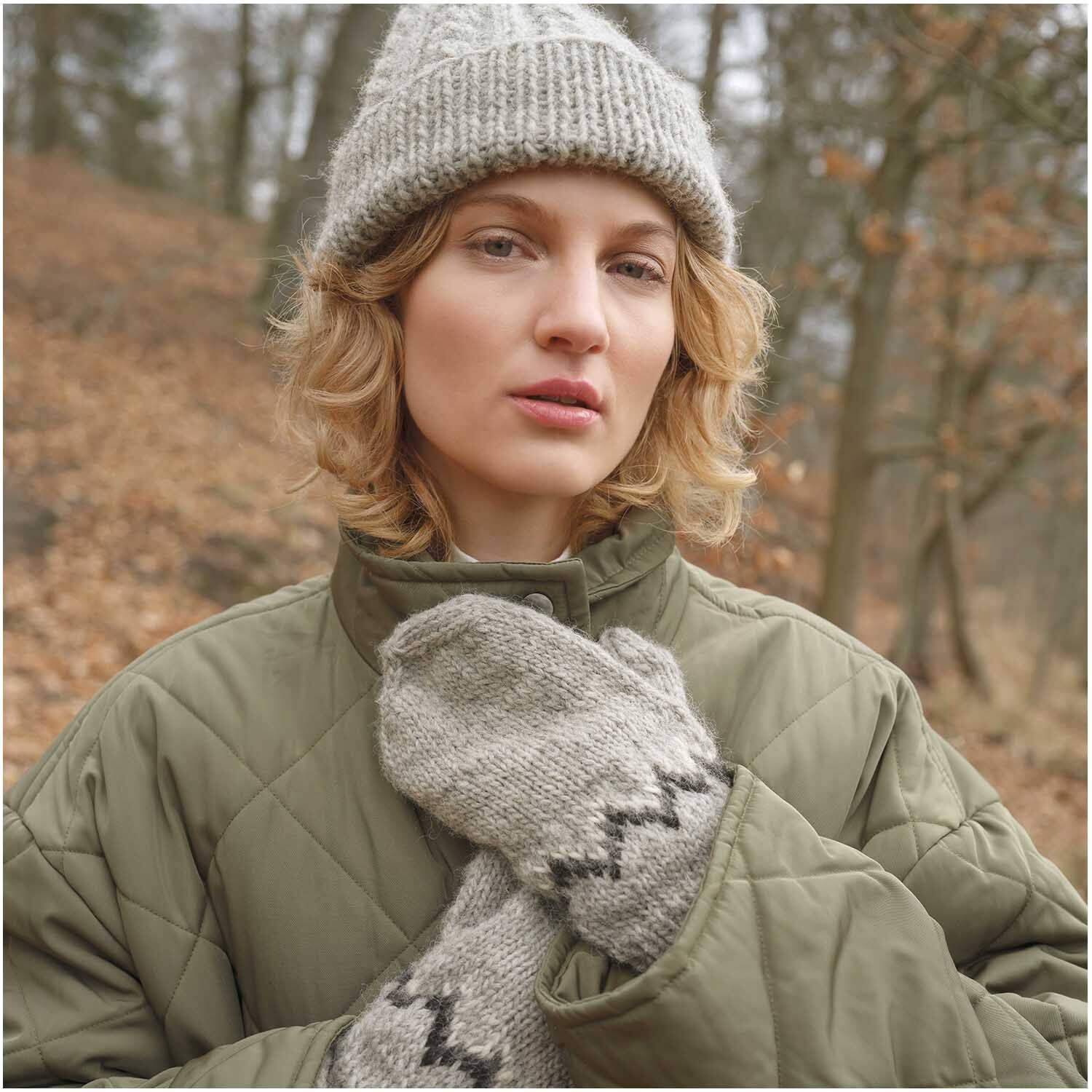 Essentials Organic Wool aran: Frau in grauer Strickmütze und Handschuhen im Wald, trägt olivgrüne Jacke. Ideal für kalte Tage.