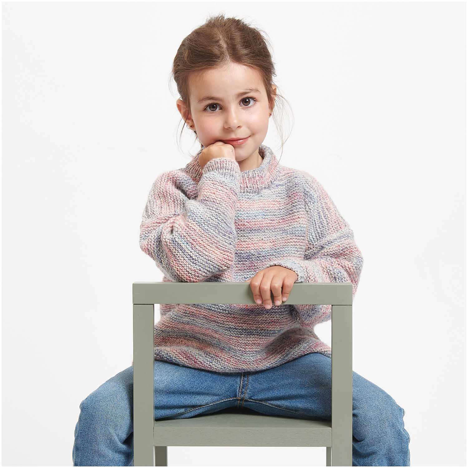Creative Colour Shift dk: Kind in gestricktem Pullover in Pastelltönen sitzt auf einem Stuhl, lächelnd.