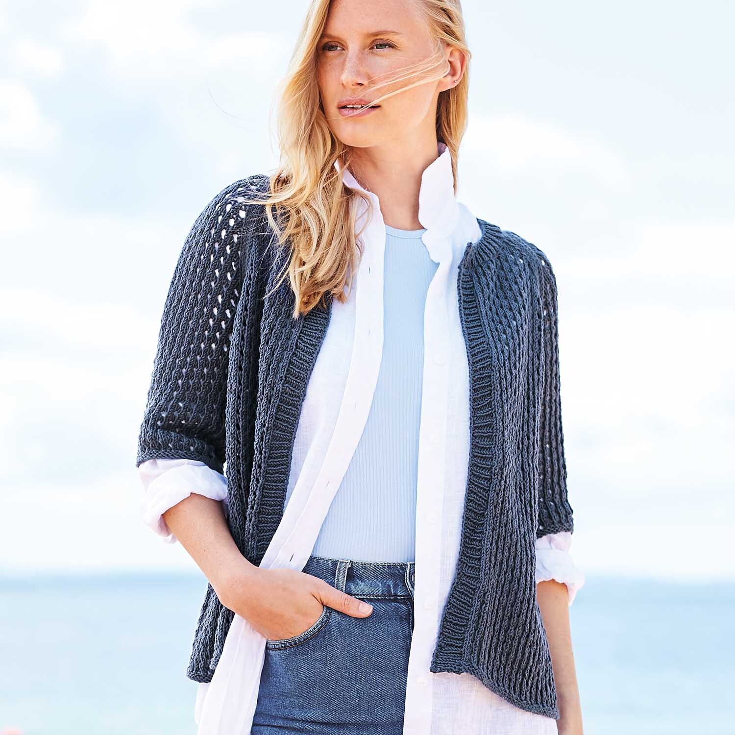 Strickanleitung Lochmuster-Jacke aus Essentials Organic Cotton aran: Frau trägt eine dunkelblaue, gestrickte Jacke über einem weißen Hemd am Strand.