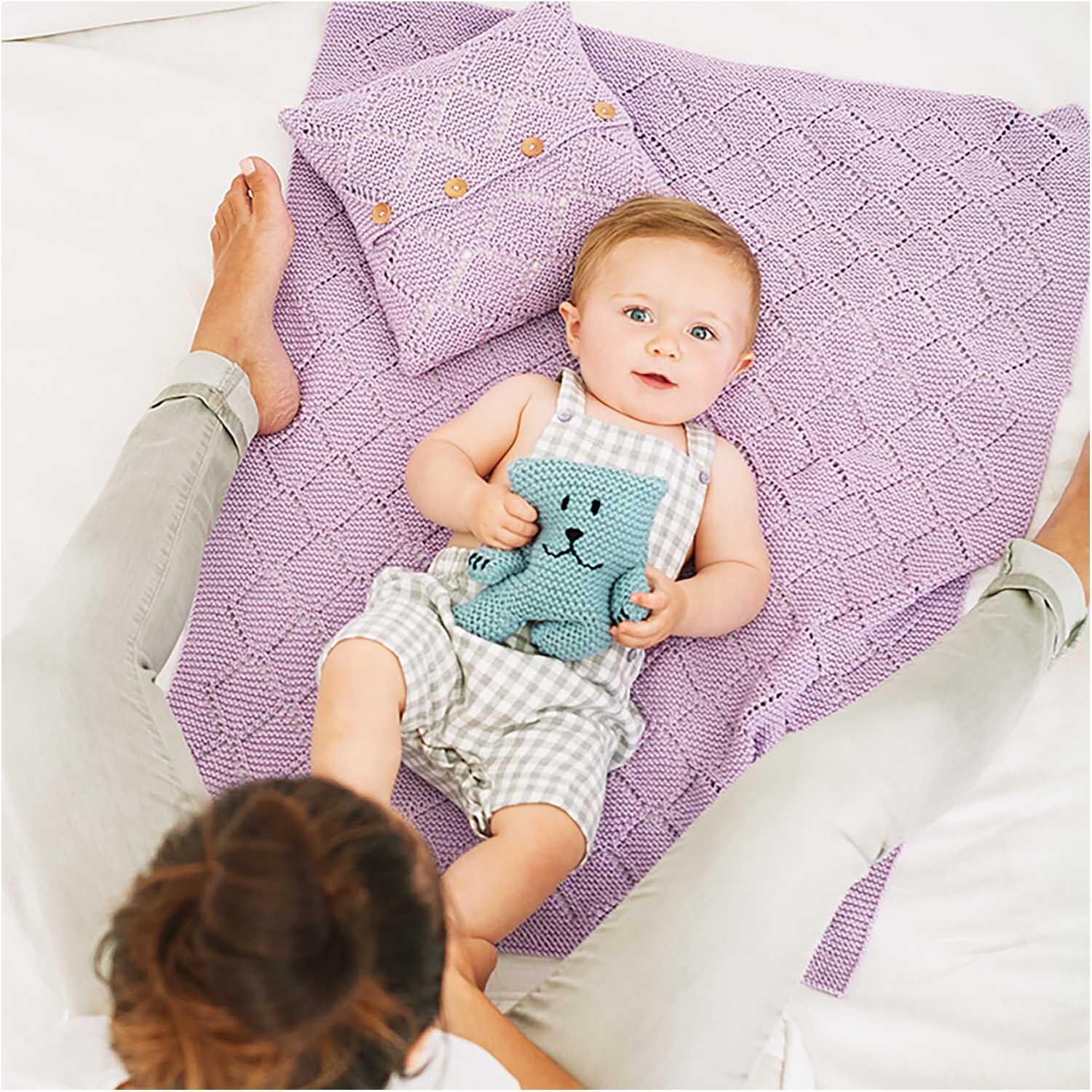 Rico Baby Cotton Soft dk: Ein Baby liegt auf einer gestrickten, lila Decke mit einem Kissen.
